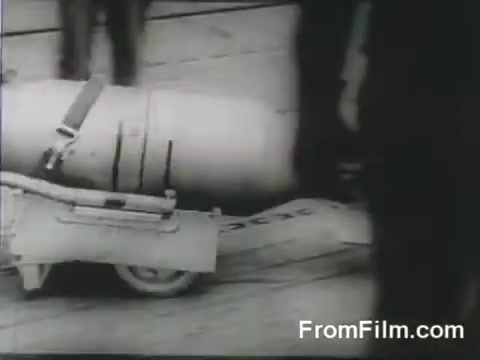 The still captures a moment during the Doolittle Raid, showing a bomb being maneuvered on a wheeled platform. The context is the preparation for the launch of B-25 bombers from the USS Hornet on April 18, 1942, highlighting the logistical efforts behind the historic air raid. The scene is likely bustling with crew members, emphasizing the urgency and tension of the mission.