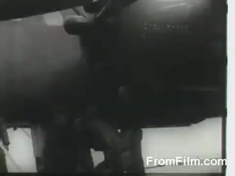 The still depicts a close-up view of a B-25 bomber, capturing the intricate details of its engine and fuselage as part of the Doolittle Raid on April 18, 1942. The shot likely emphasizes the aircraft's preparation for takeoff from the USS Hornet, showcasing the wartime technology and effort involved in this historic mission. The black-and-white format enhances the vintage feel of the footage.