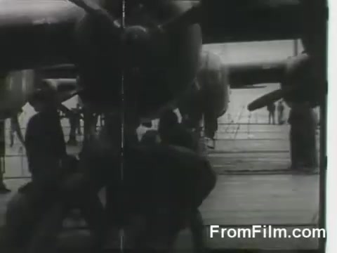 The still captures the moment of the Doolittle Raid on April 18, 1942, showcasing B-25 bombers being prepared for launch from the USS Hornet. Ground crews are seen working around the aircraft, emphasizing the urgency and teamwork involved in this historic military operation. The image is in black and white, reflecting the documentary style of the era.