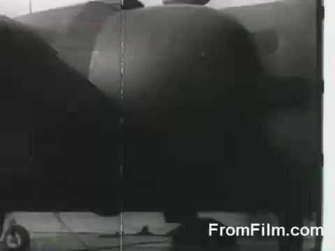 The still depicts a B-25 bomber as it is being prepared for takeoff from the USS Hornet on April 18, 1942, during the Doolittle Raid. The image captures a moment of military aviation history, showcasing the aircraft's distinctive features and the context of its launch from an aircraft carrier. The image is likely in black and white, typical of 16mm film from that era.