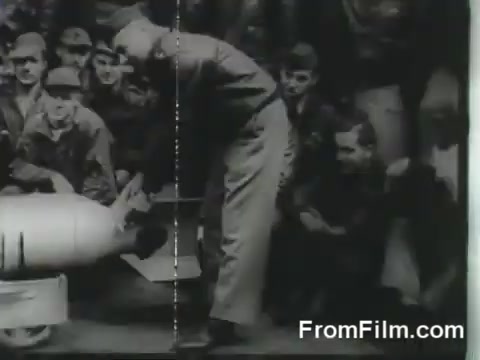 The still captures a moment from the Doolittle Raid preparation, featuring a man leaning over a bomb as a group of soldiers watch attentively. The setting appears to be on the USS Hornet, with uniforms indicating military personnel engaged in the mission. The atmosphere conveys a sense of focus and teamwork as the raiders prepare for their historic launch on April 18, 1942.