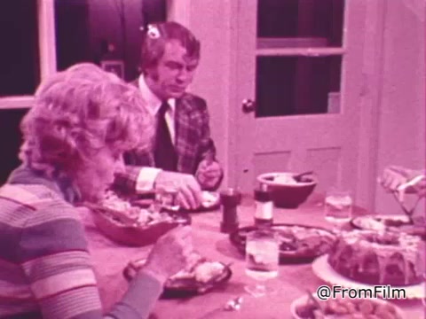 The still features a dinner scene with two people seated at a table. A woman with curly hair is eating, dressed in a colorful striped sweater, while a man in a checkered suit is focused on his meal. The table is set with various dishes and drinks, suggesting a communal meal. The atmosphere appears warm and inviting, reflective of the sharing theme in the context of the PSA.