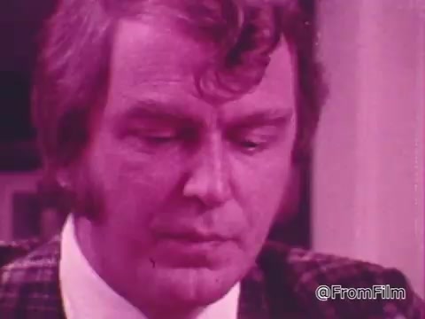 The still from 'One Great Hour of Sharing PSA' depicts a man with slightly curly hair, looking down thoughtfully. He wears a suit jacket that appears patterned, with a white shirt underneath. The image has a notable purple hue, creating a distinct visual effect. The context suggests a focus on themes of contemplation and community sharing, typical of public service announcements aimed at encouraging outreach and support.