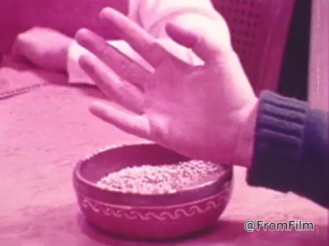 The still features a hand reaching towards a bowl filled with small objects, possibly seeds or grains, resting on a table. The scene suggests a moment of sharing or generosity, in line with the theme of the 'One Great Hour of Sharing' PSA. The overall color tone appears to be a warm, pinkish hue, contributing to an inviting atmosphere.