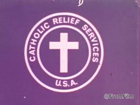 The still features the logo of Catholic Relief Services, which includes a cross centered within a circular design that has 'CATHOLIC RELIEF SERVICES' and 'U.S.A.' written around it. The background is a solid purple color, emphasizing the logo's imagery, reinforcing the organization's focus on humanitarian aid and support.