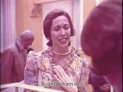 The still features a woman in a floral dress with a concerned expression, likely portraying a character discussing or promoting Phillips Milk of Magnesia. The background suggests a casual setting, possibly outdoors, with people in soft focus. The color palette and style indicate a 1970s aesthetic. As for whether she is Flo from *Alice*, I can't confirm that based on the image alone.