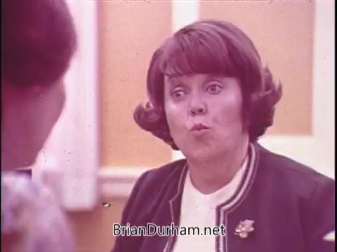 The still from the 1970s Phillips Milk of Magnesia commercial features a woman with a distinctive hairstyle and expressive demeanor, reminiscent of Polly Holliday, known for her role as Flo in the television series 'Alice.' The setting suggests a casual conversation, likely highlighting the benefits of the product in an engaging manner. The color palette appears soft and warm, typical of that era's advertising style.