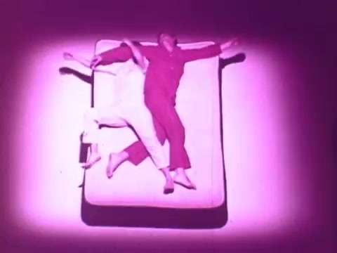 The still features a mattress set against a vibrant pink background. Two people, dressed in matching sleepwear, are playfully interacting on the mattress, showcasing comfort and support. The scene captures a light-hearted, whimsical vibe, emphasizing the relaxing qualities of the Beautyrest mattress.