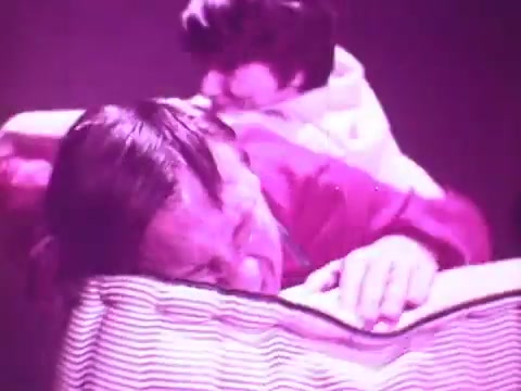The still features a man lying on a striped mattress, appearing uncomfortable, with a concerned expression. A child is positioned behind him, suggesting a playful or disruptive interaction. The lighting is vibrant purple, contributing to a surreal atmosphere typical of commercial aesthetics. Overall, it conveys a message about sleep disturbance, likely promoting the benefits of the Beautyrest mattress for restful sleep.