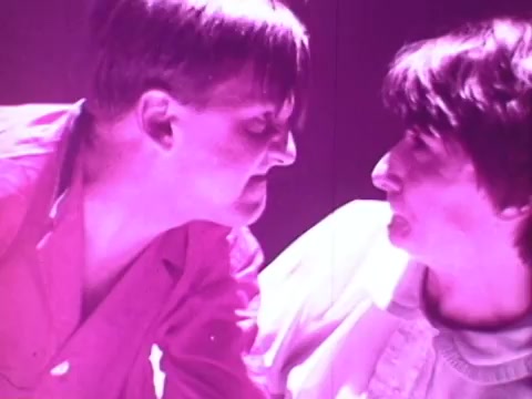 The still depicts two men in close proximity, facing each other with intense expressions. The lighting has a striking pink hue, contributing to a dramatic and surreal atmosphere. Their body language suggests a confrontation or high tension, contrasting with the typical calm associated with mattresses.