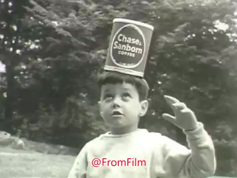 The still from the 1960s Chase & Sanborn Coffee commercial features a young boy with a can of Chase & Sanborn Coffee balanced on his head. He appears curious and slightly surprised, looking upwards. The background is blurred with greenery, suggesting an outdoor setting. The scene captures a playful moment, emphasizing the brand's legacy and connection to family, as indicated by the voice-over that references the long history of coffee roasting leading up to the present day.