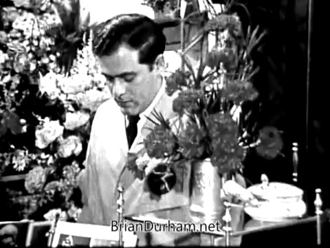 The still features a male florist in a shop surrounded by a vibrant array of flowers. He appears focused on his work, possibly arranging a bouquet. The setting suggests a classic floral shop ambiance, emphasizing the importance of flowers in celebrating occasions like graduations and Mother's Day. The commercial aims to highlight the emotional connection expressed through floral gifts, especially for those unable to attend events in person.