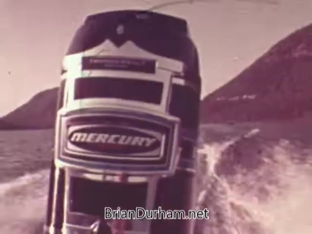 The still from the 1960s Mercury Outboard Engines commercial features a close-up view of a Mercury outboard motor mounted on a boat. The engine is prominently displayed, showcasing its branding, while water splashes behind, indicating movement. The backdrop includes mountainous terrain, suggesting an outdoor setting. The overall tone is nostalgic, highlighting the boat's speed and adventure associated with Mercury products during that era.