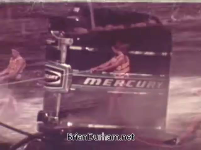 The still from the 1960s Mercury Outboard Engines commercial features a close-up shot of a Mercury outboard motor prominently displayed. Two individuals, likely water skiers, can be seen in the background. The scene conveys a sense of excitement and adventure associated with boating and water sports, highlighting the performance and reliability of Mercury outboard engines. The color palette reflects the era's vintage aesthetic.