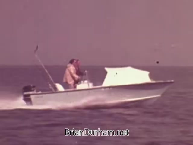 The still depicts a small boat navigating through water, powered by a Mercury outboard engine, likely filmed in the 1960s. Two individuals are visible, engaged in the experience of boating. The scene suggests leisure and adventure, typical of promotional content aimed at showcasing the utility and performance of Mercury outboard engines. The colors and styling reflect the era's aesthetic.
