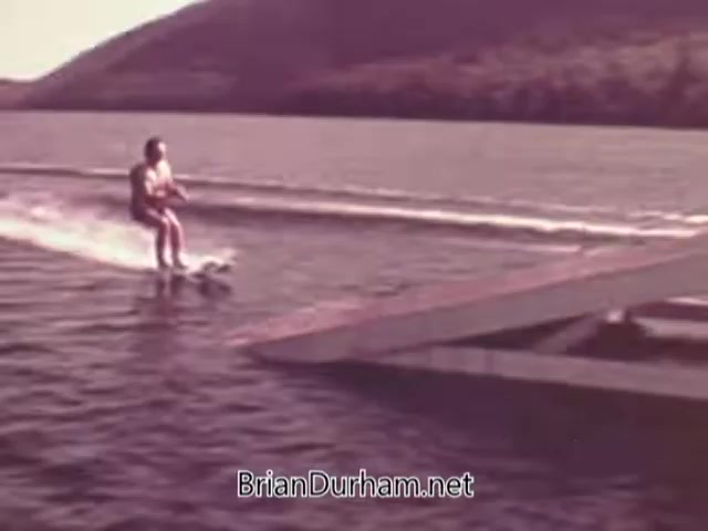 The still features a man water skiing on a lake, likely showcasing the performance of Mercury Outboard Engines from the 1960s. The scene captures a serene outdoor environment with hills in the background and a dock visible on the right. The focus is on leisure and recreational boating activities, emphasizing the speed and agility of the outboard engines.