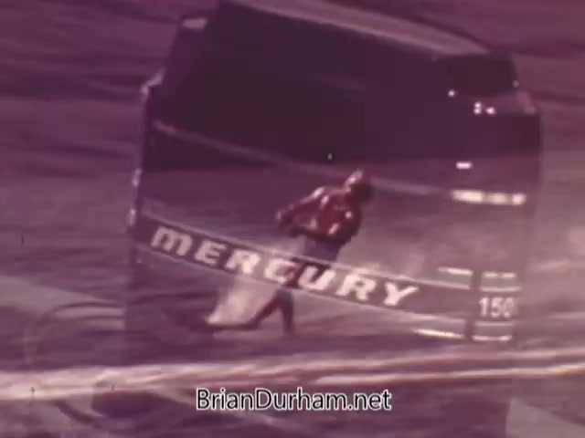The still features a dynamic scene from a 1960s Mercury Outboard Engines commercial, showcasing a powerful outboard motor. In the foreground, a water skier is captured in action, emphasizing the motor's performance and speed. The background prominently displays the 'MERCURY' logo, highlighting the brand's identity and promoting its appeal to boating enthusiasts. The overall aesthetic is vintage, with a nostalgic feel typical of that era’s advertising.