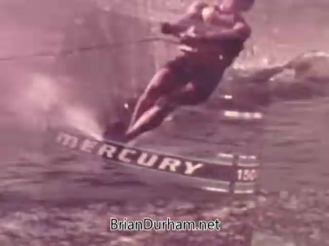 The still from the 'Mercury Outboard Engines' commercial showcases a water skier performing a dynamic maneuver on a lake. The skier is seen gripping a ski rope, with spray churning around as they navigate the water. The word 'MERCURY' is prominently displayed on the ski, emphasizing the brand's association with water sports and outboard engines. The aesthetic has a nostalgic 1960s feel, with a slightly faded color palette typical of the era's film quality.