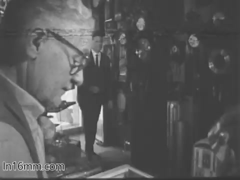 The still from the 1968 Guideposts PSA titled 'Watchmaker' shows an older man, presumably a watchmaker, focused on his work. He appears to be inspecting a watch or clock with a serious expression. In the background, a man in a suit stands observing him. The setting is filled with various clocks and watch-related items, creating a warm, nostalgic atmosphere. The black-and-white aesthetic adds to the vintage feel of the scene.