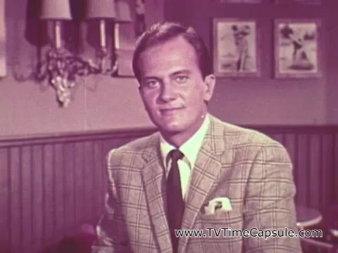 The still features Pat Boone in a well-tailored suit, addressing the camera with a friendly demeanor. He is likely in a casual setting, possibly a living room or a studio, with framed pictures on the wall. Boone is promoting U.S. Savings Bonds as a vital tool for supporting national security and the military efforts during the Vietnam War era. His message reflects the patriotic sentiment of the time, emphasizing individual contributions to the country’s efforts abroad.