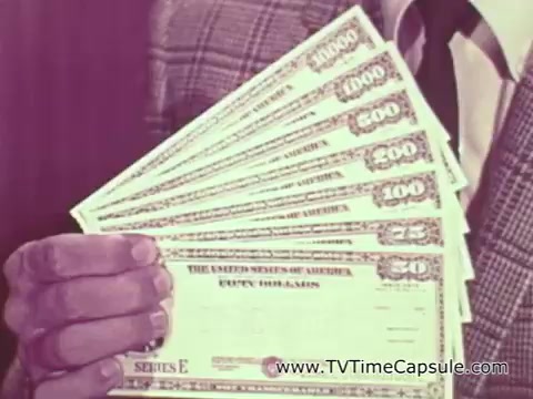 The still features a close-up of a man's hand holding several U.S. Savings Bonds, showcasing various denominations, including a $1,000 bond and smaller ones like $100 and $50. The man is dressed in a suit, and the visual emphasizes the importance of these bonds as a means of supporting national security and the military effort in Vietnam. The PSA likely aims to appeal to viewers' patriotism and encourage financial contributions to the war effort.