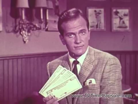 This 16mm still from the 'Pat Boone Savings Bonds PSA' features the singer and actor promoting U.S. Savings Bonds during the Vietnam War era. Boone appears in a smartly dressed outfit, holding several savings bonds, emphasizing their importance as a contribution to national security and support for American troops. The backdrop includes a wooden wall and framed pictures, creating a homey, reassuring atmosphere during a time of conflict. The message underscores the call for Americans to invest in savings bonds as a patriotic act.