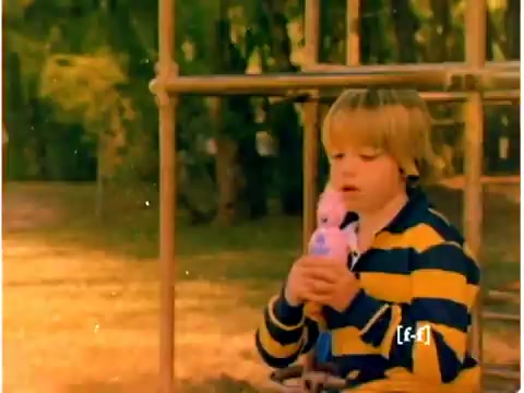 The still from the 1970s Mr. Bubble TV commercial features a young boy sitting on playground equipment. He wears a striped yellow and black sweater and is holding a pink toy, likely a character associated with the commercial. The background is filled with greenery, indicating an outdoor setting. The colors appear faded, reflecting the deterioration common in CRI film over the years. The overall atmosphere evokes a nostalgic childhood scene.