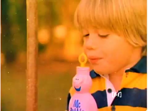 The still from the 1970s Mr. Bubble TV commercial features a young boy with blonde hair, wearing a yellow and navy striped shirt. He is interacting with a pink Mr. Bubble bottle, likely blowing bubbles. The colors appear slightly faded due to age, typical of CRI films, and the background has a warm, outdoor ambiance, possibly a park or backyard setting.