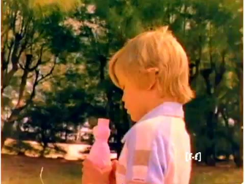 The still features a young child holding a pink bottle of Mr. Bubble. The boy has light blonde hair and wears a striped shirt with pastel colors. In the background, there are trees with vibrant green leaves, contributing to the nostalgic feel of the 1970s. The colors appear faded, typical of CRI films from that era, indicating the wear of the negative over time.