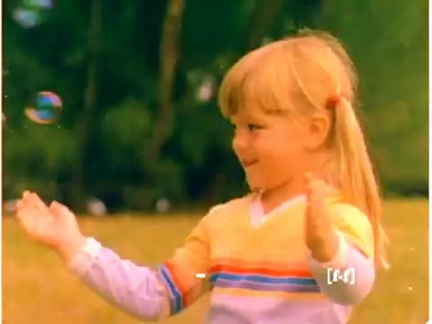 The still captures a nostalgic moment from a 1970s Mr. Bubble TV commercial, featuring a young girl in a bright, colorful sweater. She has blonde hair styled with a playful part and is joyfully interacting with bubbles in a grassy outdoor setting. The colors appear somewhat faded, typical of aging CRI film. The scene exudes a sense of innocence and fun, synonymous with childhood joy.