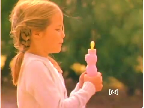 The still from the 1970s Mr. Bubble TV commercial features a young girl with a light brown braid, holding a purple bottle designed to resemble the Mr. Bubble character. She stands outdoors, softly lit, with a gentle expression. The colors have faded due to the aging of the film, contributing to a nostalgic, warm ambiance reminiscent of classic childhood moments. The overall composition captures the innocence and joy associated with bath time play.