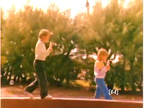 The still captures two children playing outdoors, likely enjoying a sunny day. They are blowing bubbles, with one child dressed in a white sweater and black pants, and the other wearing a pale top and blue pants. In the background, lush greenery can be seen, indicative of a playful, carefree atmosphere typical of 1970s childhood commercials. The color quality reflects a slight aging, common in CRI films, adding a nostalgic feel to the scene.