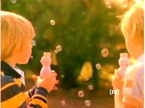 The still features two children, likely siblings, holding bubble wands near a sunny outdoor setting. One child is wearing a striped yellow and black shirt, while the other is dressed in a light outfit. Bubbles float around them, creating a playful and nostalgic atmosphere typical of 1970s commercials. The overall color quality appears faded, indicative of aging film stock. The scene captures the joy of childhood and the fun associated with Mr. Bubble products.