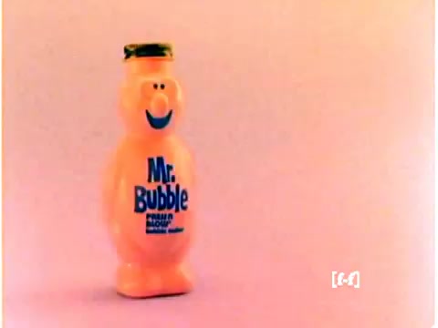 The still features a bottle of Mr. Bubble, designed with a cheerful, rounded character shape. The bottle is predominantly pink, emphasizing the playful and fun nature of the product. The label prominently displays the name 'Mr. Bubble' in blue, along with a smiling face, which adds to its appeal. The background is a soft, pastel hue, typical of 1970s aesthetic, but the colors show signs of fading due to age, characteristic of CRI film from that era. The image captures the nostalgic essence of the commercial.