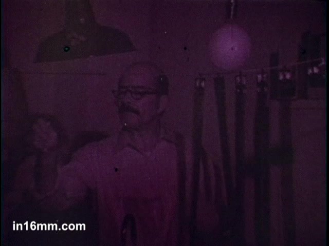 The still from the 'Ajax Cleanser' commercial shows a man with glasses in a dimly lit environment, possibly demonstrating the product. He appears engaged and may be sharing its effectiveness, as indicated by the reference to the cleansing product turning blue. There are items hanging in the background, adding to the domestic setting. The overall tone is nostalgic, characteristic of mid-century advertisements.