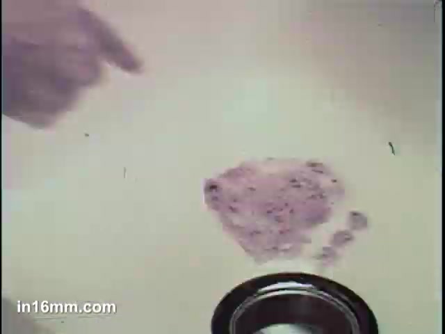 The still features a hand pointing at a pile of purple powder on a surface, likely representing Ajax Cleanser. A black container is visible nearby, suggesting the use of the product. The visual emphasizes the transformation aspect of the cleanser, tying into the 'It Turns Blue' theme, indicating a change when the product is used. The background is neutral, directing focus to the powder and action.