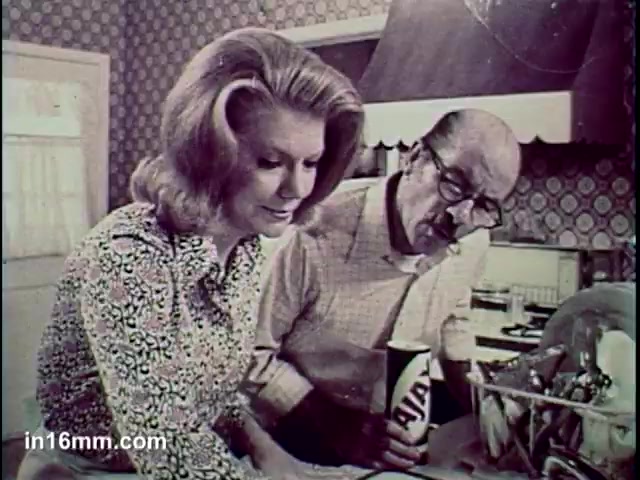 The still from the 'Ajax Cleanser - It Turns Blue' commercial features a woman and a man in a kitchen setting. The woman, wearing a patterned blouse, appears to be cleaning a surface, while the man, holding a container of Ajax Cleanser, observes her, suggesting collaboration or guidance in a household task. The background has vintage wallpaper, enhancing the retro aesthetic of the commercial.