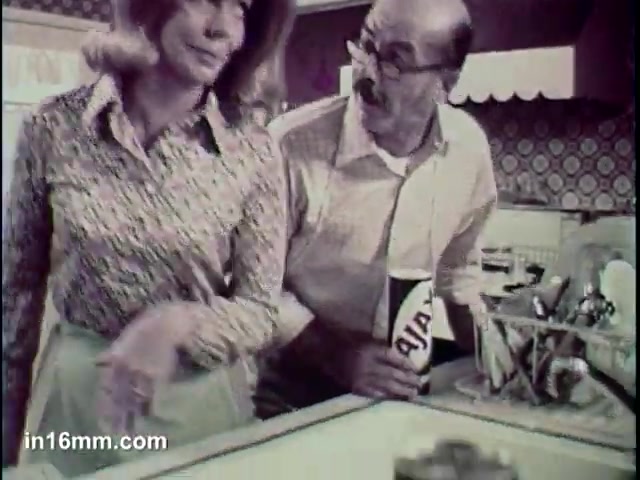 The still from the 'Ajax Cleanser' commercial features a man and a woman in a kitchen setting. The man, holding a container of Ajax cleanser, appears animated while speaking to the woman, who is dressed in a patterned blouse and looks somewhat disinterested. The background showcases typical kitchen decor of the time, including patterned wallpaper and a dish rack, emphasizing the domestic setting. The commercial promotes the effectiveness of Ajax cleanser, highlighting its ability to 'turn blue.'