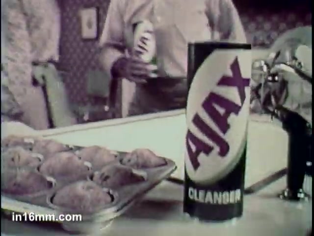The still features a kitchen setting where a large canister of Ajax Cleanser is prominently displayed. In the foreground, there is a tray of baked goods, possibly muffins, suggesting a domestic environment. A person in the background holds a can, likely another cleaning product or Ajax itself. The visual emphasizes the brand's appeal in a household setting, highlighting its effectiveness. The style reflects mid-20th century advertising aesthetics.