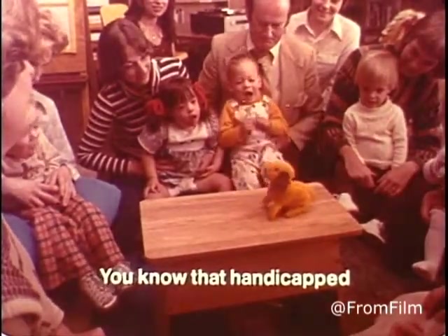 The still from the 16mm film 'Closer Look - 1970's PSA Parents' Campaign for Handicapped Children and Youth' depicts a group of children and adults gathered around a small table. At the center, a puppet or toy dog captivates the children's attention. The atmosphere seems warm and engaging, with a focus on encouraging inclusivity and understanding toward handicapped children, as conveyed by the text at the bottom: 'You know that handicapped.' The setting is likely a classroom or community space, reflecting the educational intent of the PSA. Follow @FromFilm on Twitter for more insights.