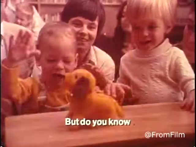 The still from 'Closer Look - 1970's PSA Parents' Campaign for Handicapped Children and Youth' features a warm scene with children interacting joyfully. A young child with light hair, wearing a yellow outfit, appears to be excited and reaching out, while another child, also light-haired, watches attentively. A woman in the background is smiling, creating a positive and engaging atmosphere. The toy in front of them resembles a small, animated dog, and the overlay text reads, 'But do you know,' hinting at an engaging message or question related to the campaign.