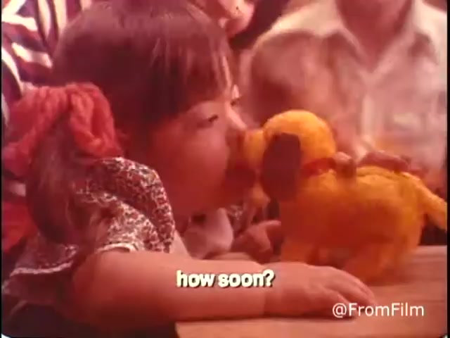 The still features a young girl interacting with a plush toy dog, seemingly engaged and curious. The caption reads 'how soon?' suggesting a focus on communication or anticipation. The warm colors and the child's expression evoke a sense of innocence and interest, typical of the messaging in the '70s PSA aimed at raising awareness for handicapped children and youth. The context emphasizes empathy and understanding in a community setting.