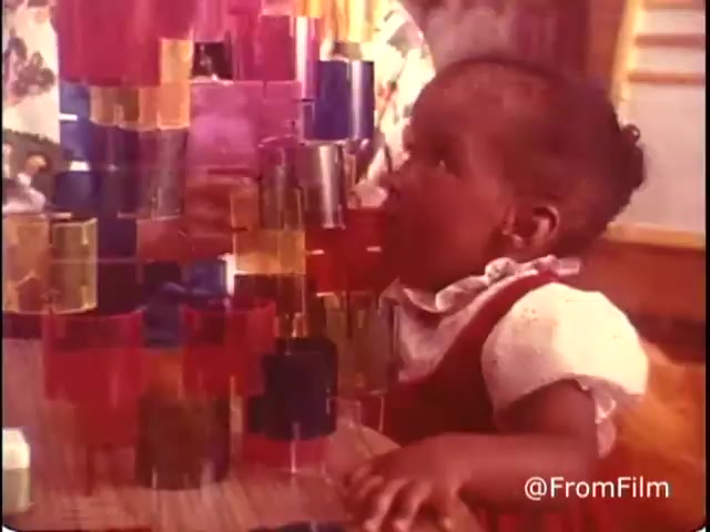 The still captures a young child engaged with colorful building blocks, concentrating on stacking them. The child is wearing a red dress with a white collar, and the vibrant blocks are a mix of colors translucent and solid. There's an adult figure in the background, suggesting a supportive environment. The image emphasizes play and creativity, reflecting the campaign's message of inclusion and interaction for children with disabilities.