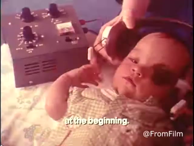 The still depicts a young child wearing headphones connected to a device that appears to be used for audio testing or therapy. The child looks curious and relaxed, with one hand raised. The background suggests a caring environment, possibly a clinic or home setting. The text at the bottom, 'at the beginning,' indicates an initial phase of a process, likely referring to early intervention strategies for children with disabilities. This PSA emphasizes the importance of support for handicapped children and youth.