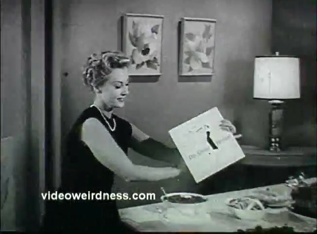 The still from the 1950's Kaiser Broiler Aluminum Foil Commercial features a woman in a black dress and pearls, demonstrating cooking techniques. She holds a card, likely showcasing a recipe or instructions, while a prepared meal is visible on the table. The setting includes floral artwork and a lamp, emphasizing a domestic and festive atmosphere ideal for Thanksgiving. The scene captures the era's emphasis on convenience in meal preparation using aluminum foil.