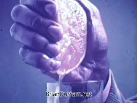 The still features a close-up of a hand squeezing an orange, with juice flowing into a glass below. The background has a subtle blue tint, highlighting the freshness of the orange juice. The focus on the action emphasizes the product's natural qualities.