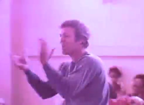 The still depicts a man in a casual sweater, animatedly gesturing as he speaks or leads a discussion in a group setting. The lighting has a pink hue, creating a warm, intimate atmosphere. In the background, a few individuals appear engaged, highlighting a sense of community. The context emphasizes the importance of taking blood pressure medication for the sake of loved ones, underscoring a caring message.