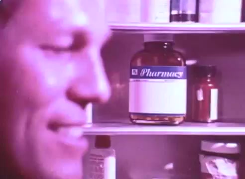 The still features a close-up of a man's profile smiling slightly, with a focus on a medicine cabinet behind him. Prominently displayed is a large bottle labeled 'Pharmas' alongside various other smaller containers. The overall tone suggests a message about the importance of taking blood pressure medication for the sake of loved ones, aligning with the campaign's theme of responsibility and care. The color palette appears warm, likely evoking a sense of comfort and encouragement.