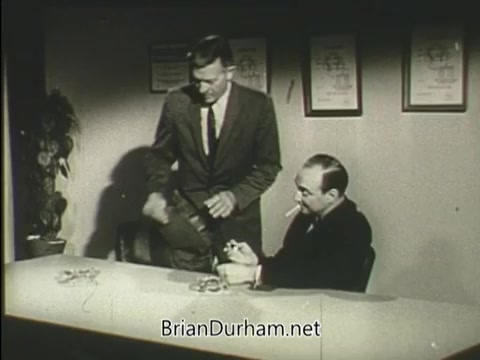 The still features two men in a vintage office setting. One man, dressed in a suit, stands beside a seated man who is holding a cigarette. The standing man appears to be interacting with an object or device, possibly related to health or medical awareness, as suggested by the context of a public service announcement. The room is decorated with framed documents or certificates on the wall, indicating a formal environment.