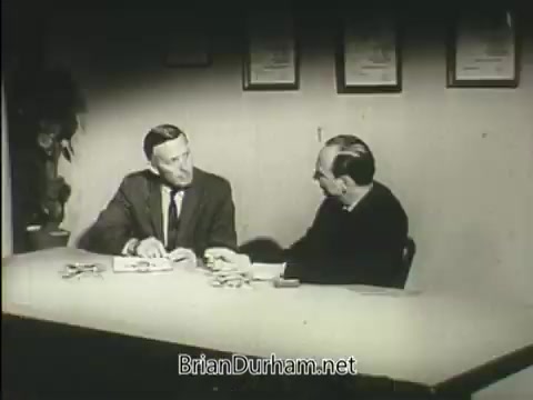 The still from the 'American Heart Association PSA' depicts two men sitting at a table. One man, dressed in a suit, is engaged in conversation, while the other seems to be listening attentively. The background features framed certificates or awards, suggesting a formal setting. The overall mood appears serious, likely reflecting the educational nature of the public service announcement regarding heart health.